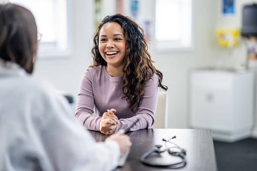 A young woman of mixed race, sits with her female doctor as they discuss her health concerns.  The patient is dressed casually and has a smile on her face.