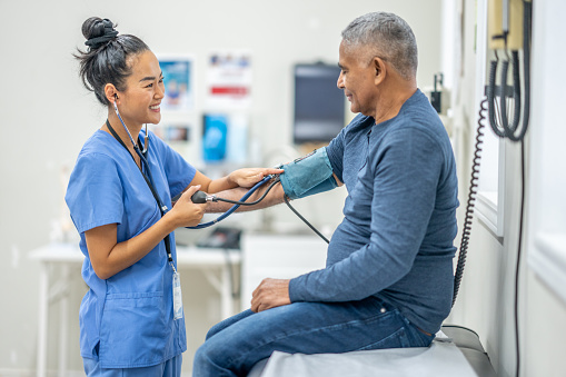 A senior gentleman of Hispanic decent, sits up on an exam table as his female nurse takes his blood pressure. The nurse is wearing blue scrubs and making small talk to put the gentleman at ease.
