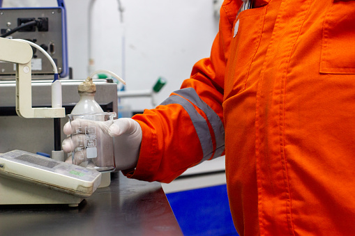 A scientist or laboratory worker wearing a bright orange protective suit and gloves is holding a beaker containing a liquid sample. The worker is conducting scientific analysis and research,