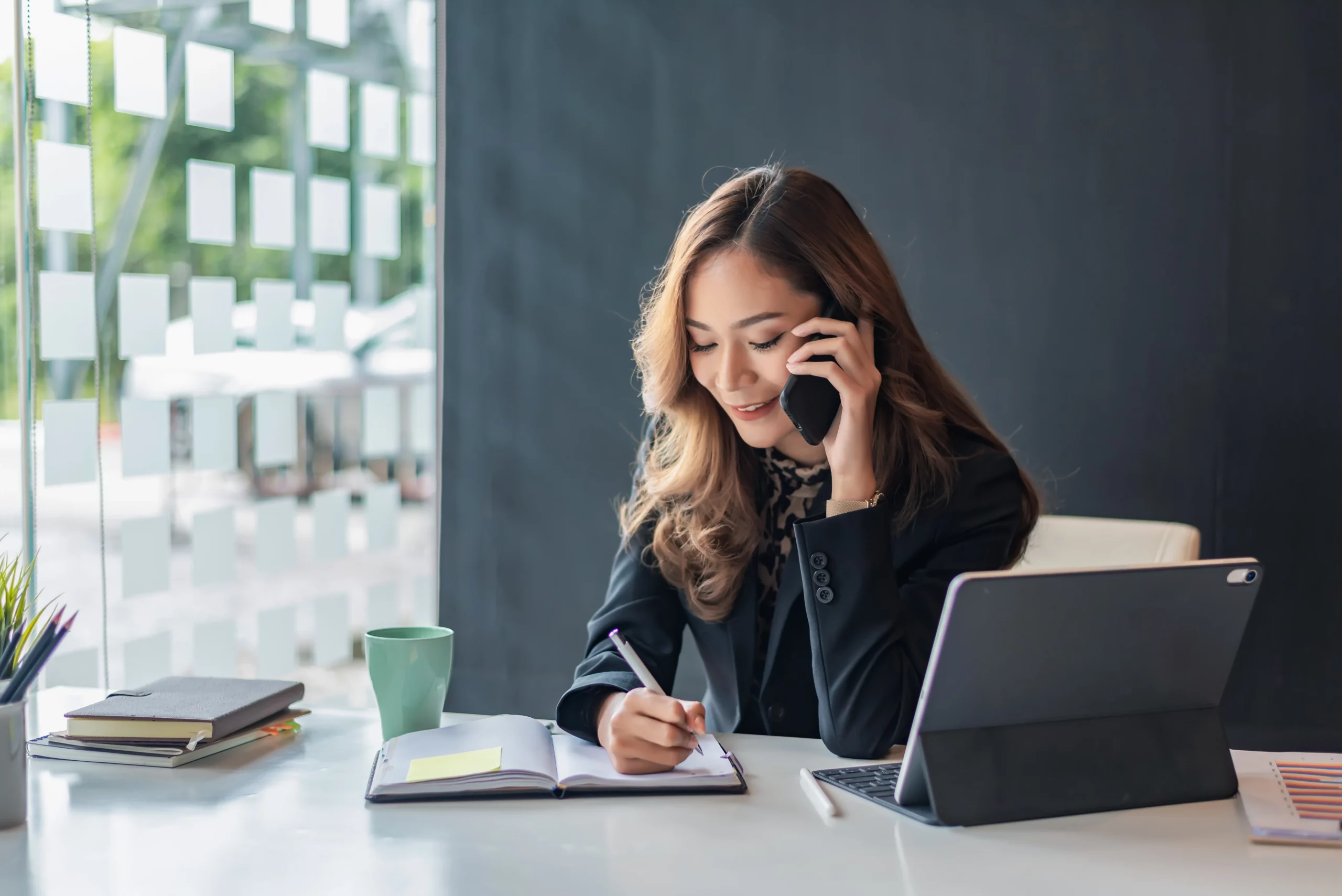 A woman in a business suit sits at a desk, talking on the phone with a focused expression.
