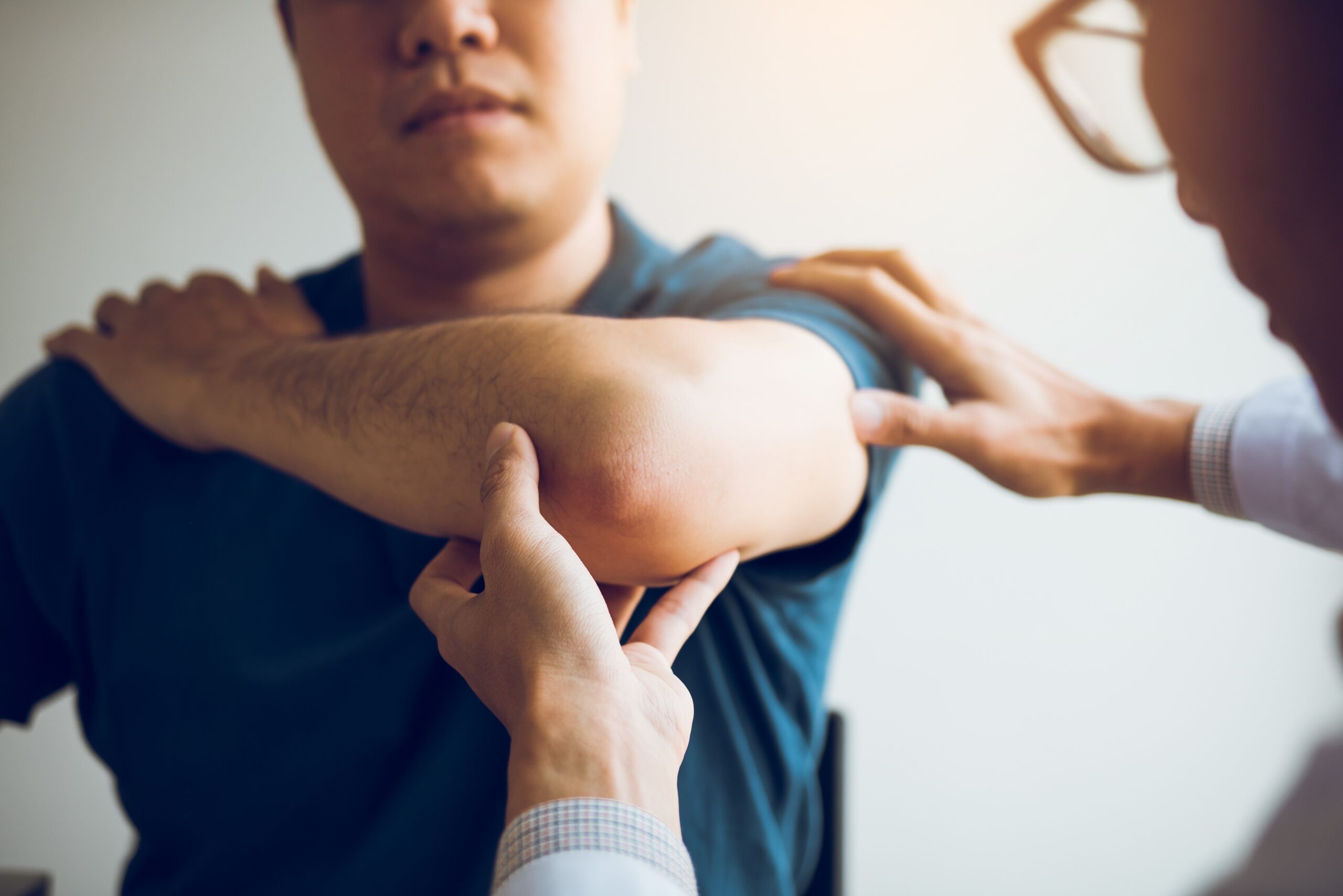 A doctor examines a patient's sore elbow, gently pressing it. The patient looks concerned. The setting is bright and clinical, suggesting a medical consultation.