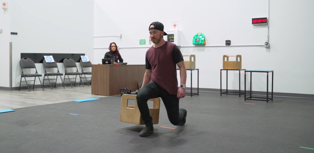 A man in a cap and casual wear performs a lunge while holding a wooden box in a spacious room. A seated woman observes in the background.