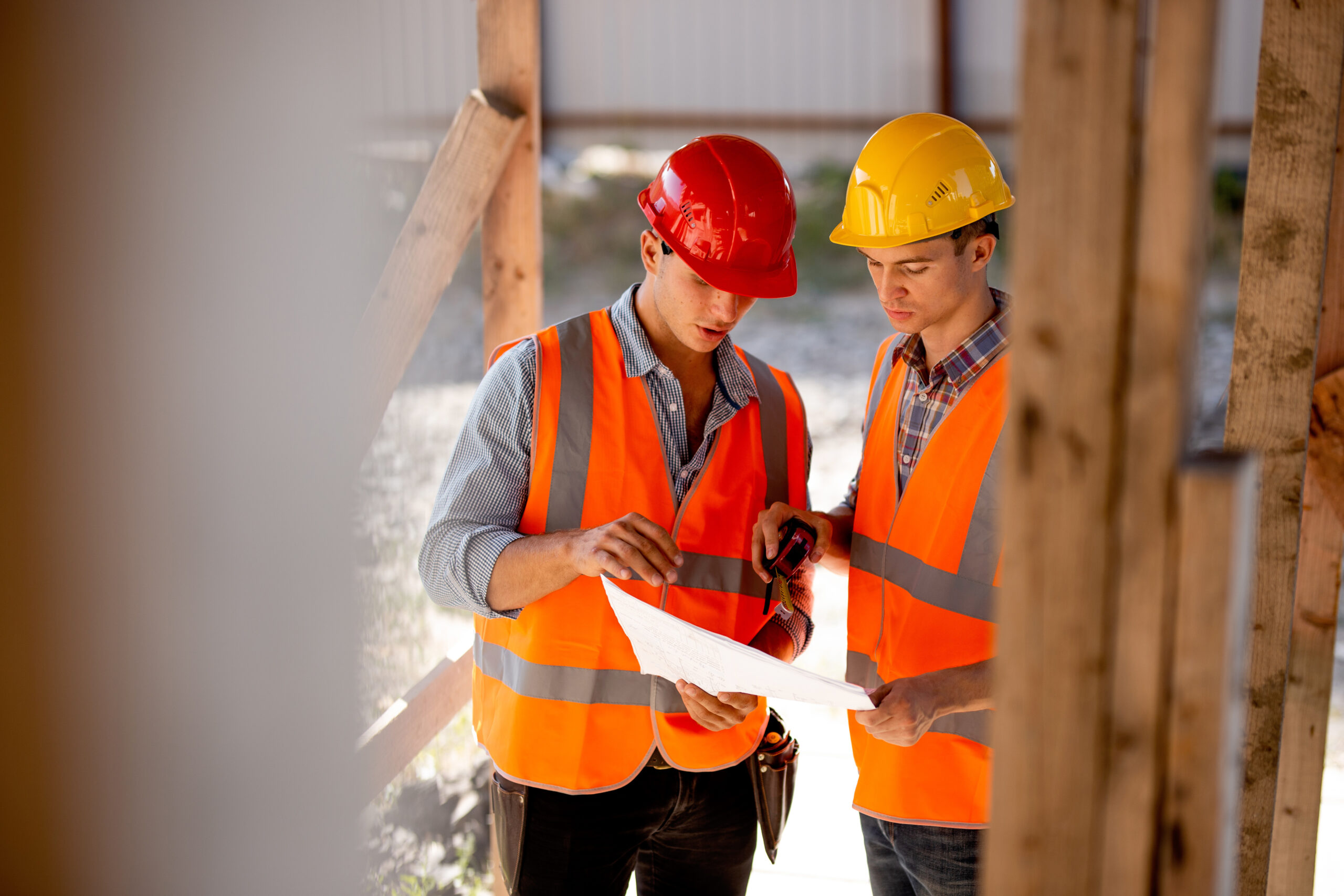 Two construction workers wearing safety vests and helmets review blueprints together at a building site.