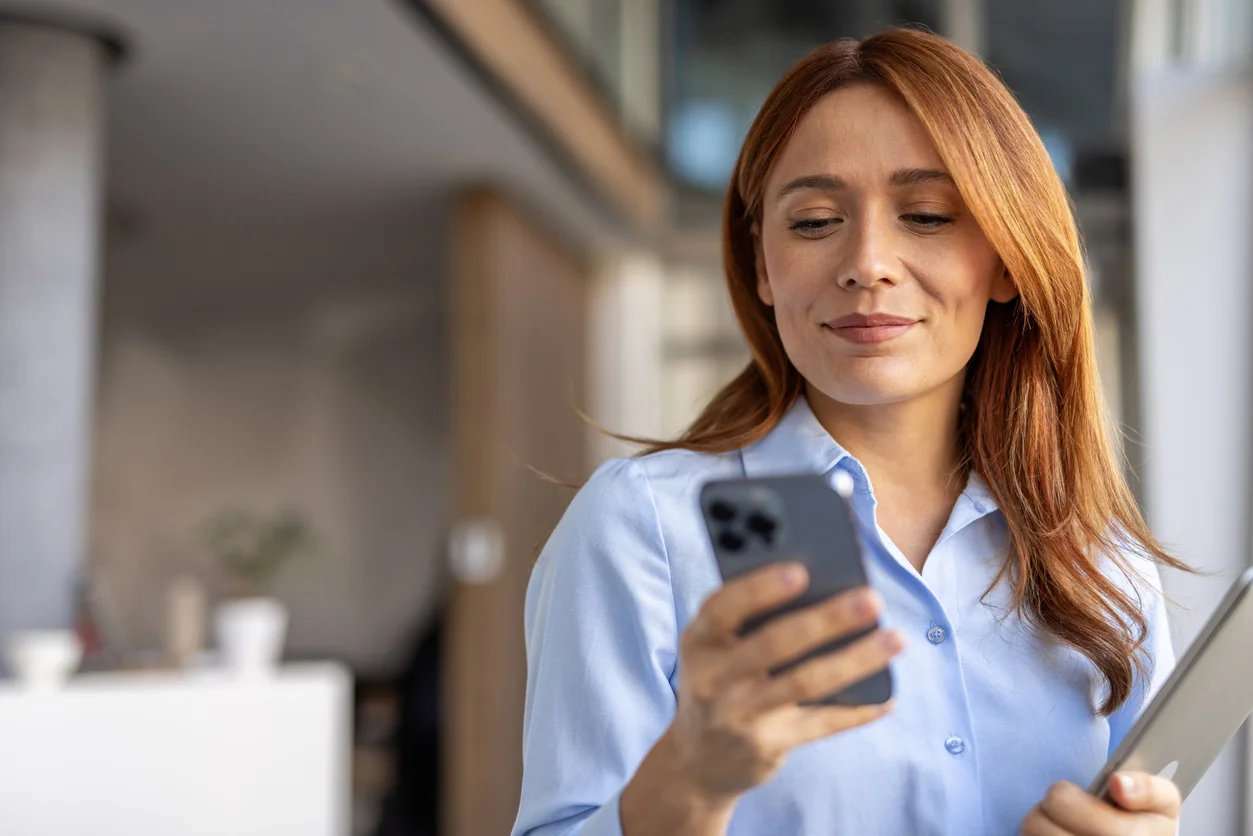 A business woman with a confident smile is standing in a modern office space, surrounded by an elegant interior. Her appearance exudes professionalism and determination, while holding a laptop and using a mobile phone. The atmosphere of the photograph emphasizes her influence and role as a leader in the modern business world.